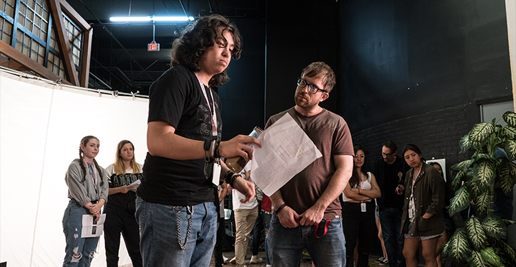 Students gather on a film set as an instructor reviews a script with a performer during a collaborative production exercise.